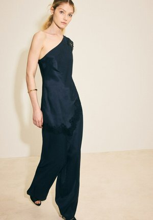 Mujer con mono de satén azul marino de un solo hombro con detalles de encaje, llevando una pulsera de oro, posando frente a un fondo beige liso.