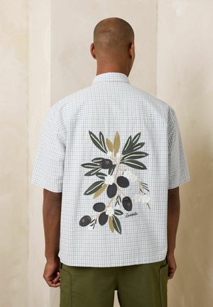 Mand iført kortærmet hvid ternet skjorte med broderede sorte oliven og grønne blade på ryggen, kombineret med olivengrønne bukser.