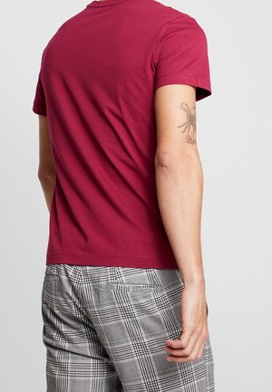 Mann trägt ein weinrotes T-Shirt mit kurzen Ärmeln und graue karierte Hosen, zeigt ein Spinnentattoo auf seinem oberen rechten Arm.