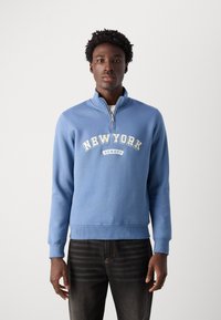 Modrá mikina s vysokým límcem, zipovým zapínáním a potiskem "NEW YORK" v krémové barvě. Navržena s žebrovaným lemem a manžetami, vyrobena z měkké látky.
