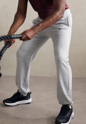 Personne portant un pantalon de survêtement gris Champion et des baskets noires, tenant et balançant des cordes de combat noires pendant l'entraînement.