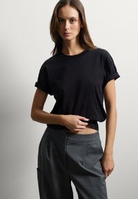 T-shirt nera in cotone con maniche corte arrotolate, abbinata a pantaloni grigi su misura con vestibilità rilassata e tasche laterali.