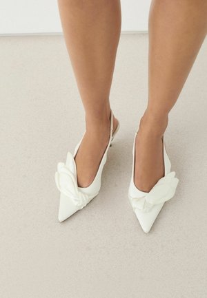 Fehér, hegyes orrú, hátul pántos magas sarkú cipőt viselő lábak, melyeken nagy, szirom alakú díszek vannak a tetején, világos padlón állnak.