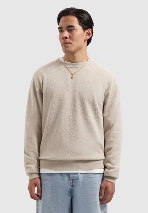 Man wearing beige textured sweater, light blue jeans, and gold necklace, standing against plain light gray background.