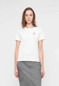 Λευκό βαμβακερό t-shirt με κοντά μανίκια, που διαθέτει ένα μικρό ροζ γραφικό λογότυπο στο στήθος, συνδυασμένο με μια γκρι φούστα.