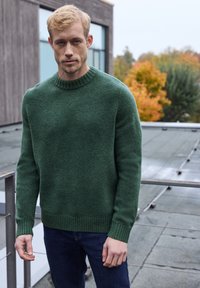 Suéter de lana verde con un cuello redondo y puños acanalados, que presenta una textura suave y un corte holgado, combinado con unos jeans azul oscuro.