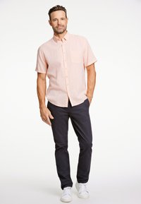 Maglietta a maniche corte rosa chiaro con bottoni e taschino sul petto, abbinata a pantaloni blu scuro e sneaker bianche, su uno sfondo neutro.