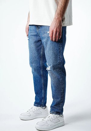 Mand iført blå revnede jeans og hvide sneakers, stående mod en ensfarvet hvid baggrund.