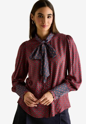 Red blouse with blue accents, featuring a bow tie collar, patterned fabric, long puff sleeves, and button-down front.