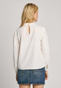 Blusa bianca a maniche lunghe con apertura a forma di chiave sul retro, tessuto lavorato e dettagli in pizzo ai polsini, abbinata a una gonna mini di jeans.