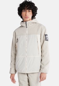 Helle beigefarbene Jacke mit Kapuze, Reißverschluss vorne und Seitentaschen. Verfügt über ein schwarzes Logo-Patch auf dem linken Ärmel und elastische Bündchen. Glatte Textur.