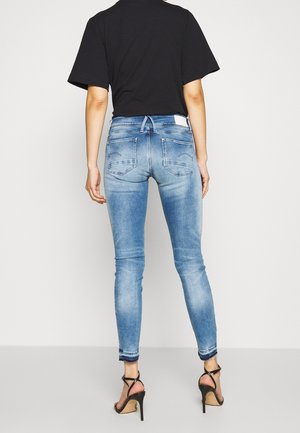 Persoon die lichtblauwe skinny jeans draagt met rafelige zomen, een zwart topje met korte mouwen en zwarte sandalen met hoge hakken, gezien van achteren.