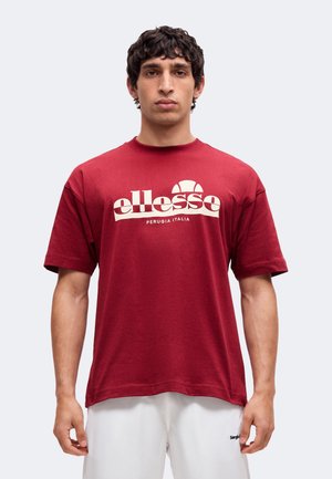 Man with short curly hair wearing a red ellesse t-shirt and white shorts, standing against a plain light background.