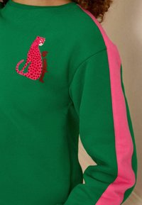 Felpa verde con strisce laterali rosa. Grafica in evidenza con stampa leopardata rosa e testo "le weekend" sul petto. Tessuto morbido.