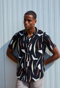 Chemise noire à manches courtes avec des motifs abstraits blancs, oranges et jaunes, fermeture par boutons à l'avant, et un col décontracté. Tissu opaque.