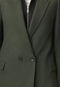 Blazer a spina di pesce verde scuro con due bottoni neri indossato sopra un indumento nero con zip e tirazip argentato.