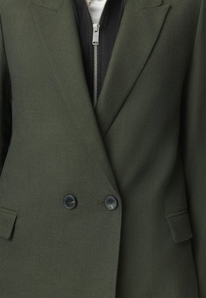 Blazer en chevrons vert foncé avec deux boutons noirs porté sur un vêtement zippé noir avec tirette en argent.
