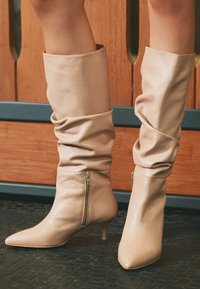 Bote negre din piele, slouchy, de lungime medie, cu fermoare laterale și vârfuri ascuțite, purtate pe un podea texturată neagră, în fața unui fundal din panouri din lemn.