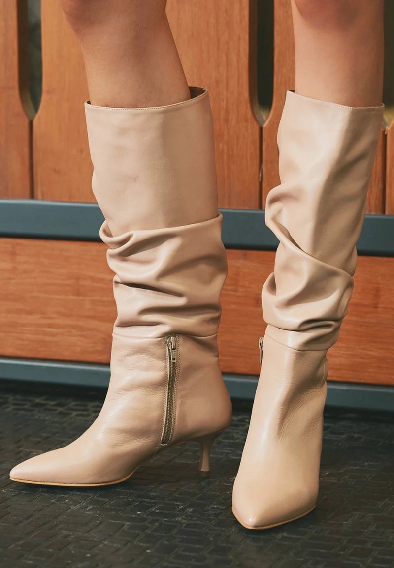 Bote negre din piele, slouchy, de lungime medie, cu fermoare laterale și vârfuri ascuțite, purtate pe un podea texturată neagră, în fața unui fundal din panouri din lemn.