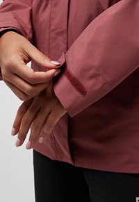 Veste imperméable bordeaux avec réglage des poignets par Velcro. Présente le logo "Texapore" sur la manche. Texture lisse, coupe ajustée.