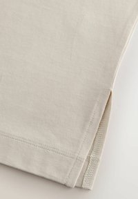 Beige bomullströja med korta ärmar, med en sid slits och kontrasterande sömmar längs fållen. Slät textur och minimal design.
