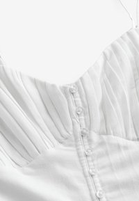 Valkoinen mekko, jossa on pleated-rakenne, nappikiinnitys takana ja ohuet säädettävät hihnat. Pehmeä kangas, jossa on sileä pinta.