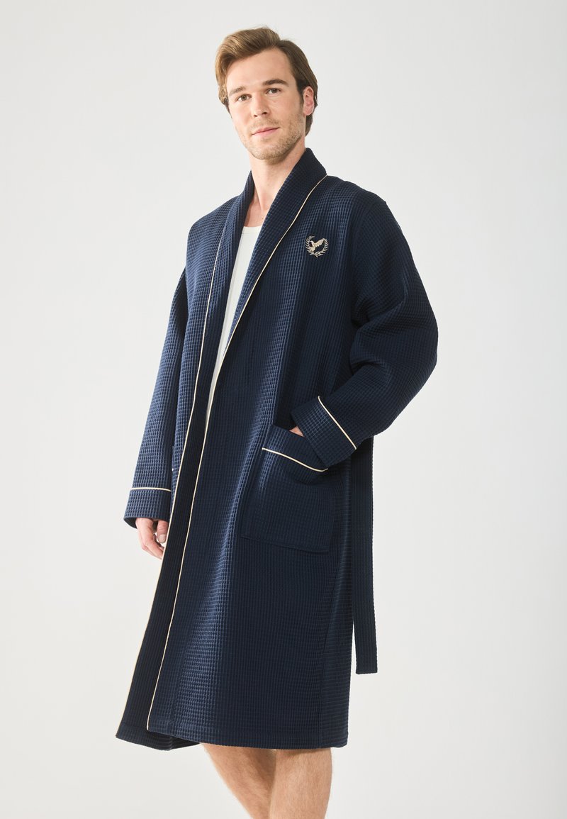 Marineblauer Bademantel mit Waffelstruktur, Schalkragen, cremefarbener Paspel und gesticktem Logo. Verfügt über zwei vordere Taschen und einen selbstbindenden Gürtel.
