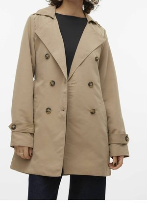 Femme portant un trench-coat beige à double boutonnage avec des boutons foncés sur un top noir et un pantalon foncé, debout devant un fond uni.
