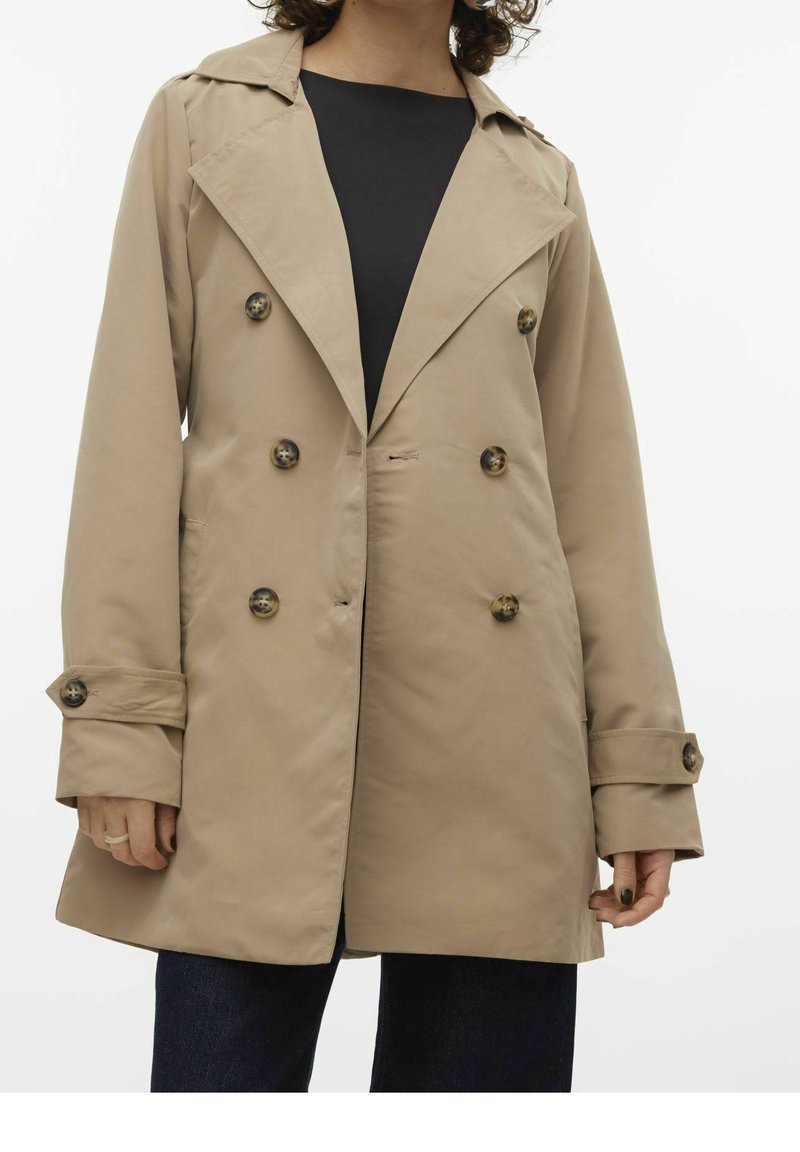 Femme portant un trench-coat beige à double boutonnage avec des boutons foncés sur un top noir et un pantalon foncé, debout devant un fond uni.
