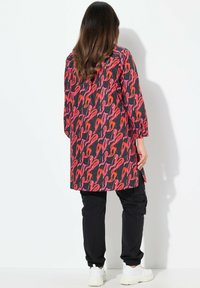 Tunica lunga in tessuto nero con un motivo astratto rosso e rosa, abbinata a pantaloni cargo neri e sneakers bianche.