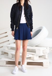 Giacca bomber navy con zipper dorato, t-shirt bianca e gonna navy plissettata. Completato con sneakers bianche. Sullo sfondo ci sono pallet bianchi impilati e pneumatici.