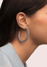 Pendientes de aro con un acabado plateado texturizado, con un diseño grueso y curvado que refleja la luz, exhibidos en la oreja de una modelo.