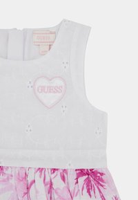 Abito bianco con motivo floreale ricamato, caratterizzato da un cuore rosa con il testo "GUESS" e una gonna con stampa floreale rosa. Chiusura con zip sul retro.