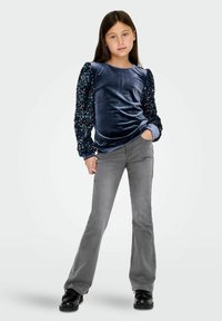 Top de terciopelo azul marino de manga larga con mangas abullonadas adornadas con lentejuelas, combinado con pantalones grises acampanados y zapatos negros, posando frente a un fondo liso.