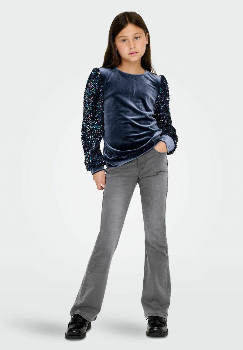 Top de terciopelo azul marino de manga larga con mangas abullonadas adornadas con lentejuelas, combinado con pantalones grises acampanados y zapatos negros, posando frente a un fondo liso.