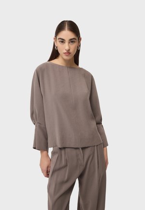 Kobieta o długich brązowych włosach, nosząca srebrne kolczyki koła oraz luźną taupe bluzkę z długim rękawem i pasujące plisowane spodnie, stoi na białym tle.