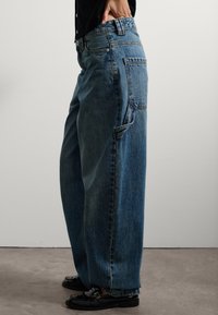 Persona con pantalones vaqueros azules de corte holgado y un bolsillo lateral para herramientas, y mocasines negros, de pie contra una pared blanca.