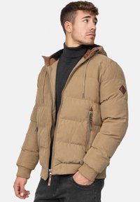Chaqueta acolchada marrón con capucha, que presenta un diseño acolchado, puños acanalados y dos bolsillos con cremallera. Tejido ligeramente texturizado con detalles en marrón.