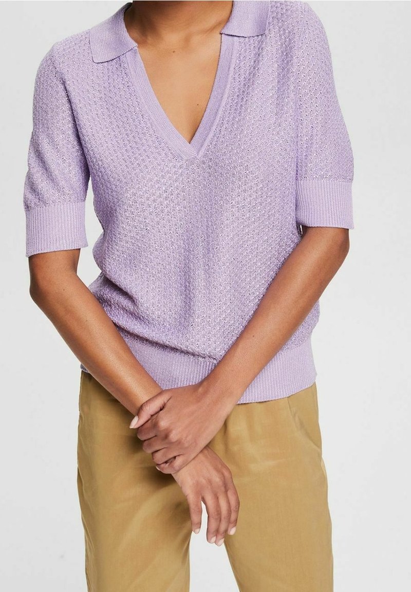 Personne portant un haut en maille lavande à manches courtes avec col en V et un pantalon beige, les bras croisés à la taille sur un fond uni.