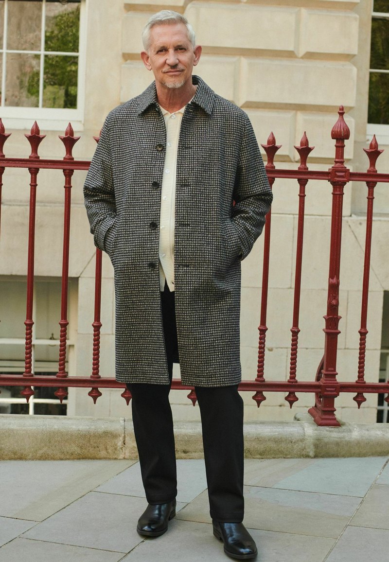 Palton gri cu model houndstooth, cu guler, închidere cu nasturi și buzunare laterale, asortat cu pantaloni negri și cizme din piele, pe un fundal cu o balustradă roșie.