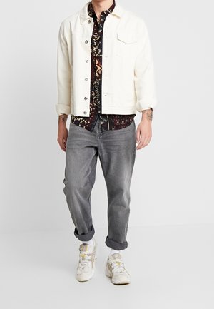 Homme portant une veste blanche boutonnée par-dessus une chemise à motifs, jean gris retroussé, baskets blanches, debout devant un fond uni.