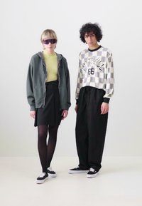 Two models stand against a plain background. One wears a grey hoodie, green top, black skirt, and black tights. The other has a checkered top and black pants.