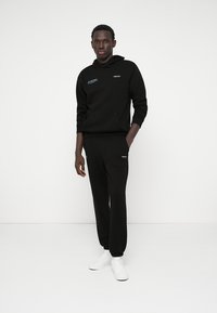 Czarna bluza z kapturem i dresy wykonane z miękkiego materiału. Bluza posiada przednią kieszeń i niebieskie logo. Spodnie slim-fit z ściągaczami przy kostkach. Białe sneakersy.