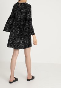 Vestido negro con patrón blanco; mangas largas y acampanadas; corsé ajustado; falda suelta; detalle de escote en forma de lágrima en la parte posterior; llevado con zapatos negros planos.