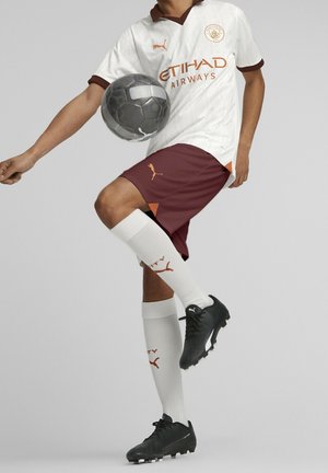 Weißes Fußballtrikot mit kastanienbraunen Akzenten, kombiniert mit kastanienbraunen Shorts. Schwarze Stollenschuhe, weiße Socken mit orangefarbenem Logo und ein Fußball.