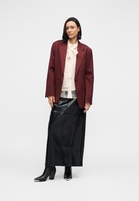 Bordeaux blazer met brede revers, crèmekleurige blouse met kanten details, zwarte leren maxirok en zwarte laarzen met hoge hak en metalen accenten.