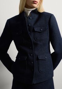 Giacca blu navy con texture, colletto alto, tasche frontali e chiusura a bottoni. Realizzata in tessuto intrecciato, con bottoni abbinati.