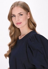 Marineblaue Bluse mit kurzen Ärmeln und gepufften Schultern, mit Rundhalsausschnitt und glatter Textur. Das Model zeigt dezent glänzende Lippen und welliges Haar.