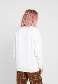 Sudadera blanca de manga larga, con puños y dobladillo de canalé, textura suave. Combinada con pantalones de cuadros marrones. Vista desde la parte trasera.