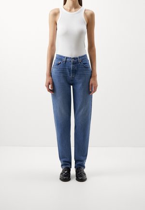 Kvinde iført hvid ærmeløs top, blå højttaljede jeans og sorte læderloafers, stående mod en ensfarvet hvid baggrund.